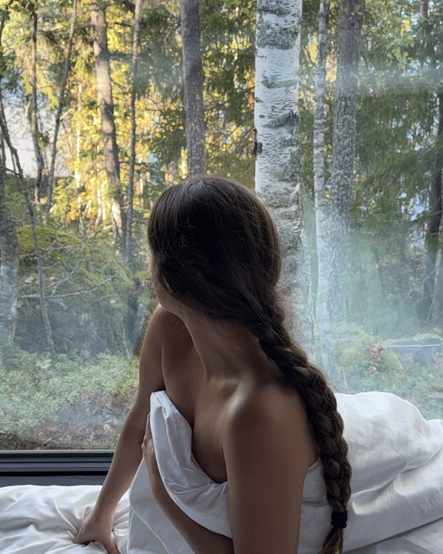 Mujer envuelta en una sábana blanca mirando al bosque por un ventanal
